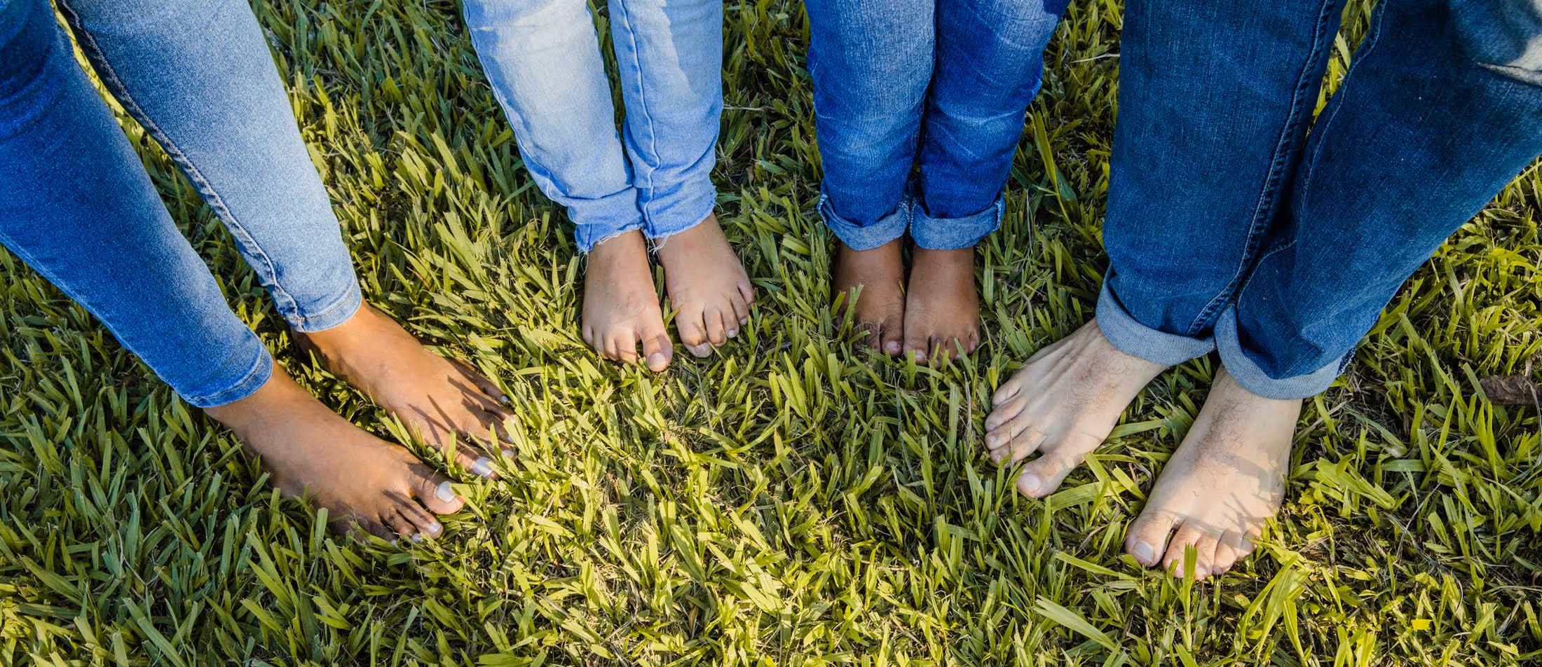 Four people standing barefoot on grass, visible from the knees down, wearing blue jeans.